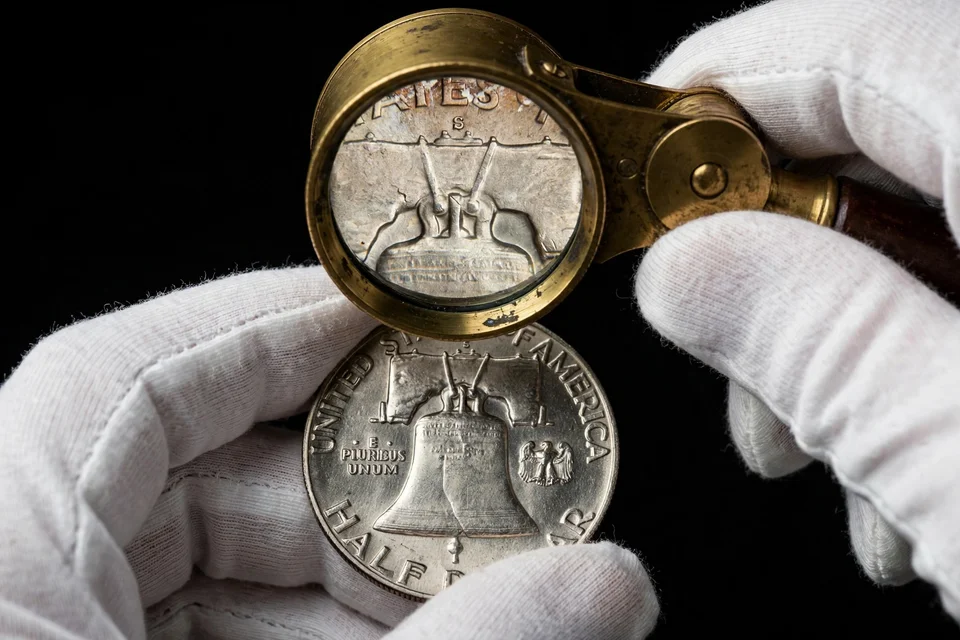Franklin half dollar reverse held in white cotton gloves under a brass loupe — mint mark above the bell